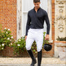 A man in equestrian attire, including a black long-sleeve shirt, white breeches, and black riding boots, walks down stone steps holding a black riding helmet in front of a brick building.