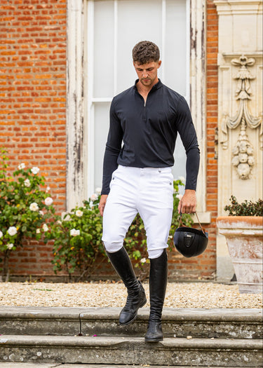 A man in equestrian attire, including a black long-sleeve shirt, white breeches, and black riding boots, walks down stone steps holding a black riding helmet in front of a brick building.
