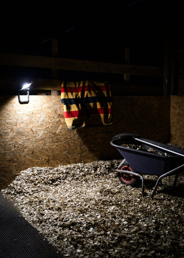 A horse stall interior at night with a wall-mounted LED light illuminating wood shavings, a striped blanket hanging on the wall, and a wheelbarrow on the right.