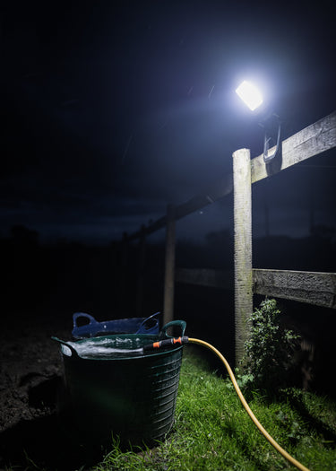 A bright outdoor floodlight mounted on a wooden fence post at night, illuminating a green bucket and a yellow hose on grass beside a fence.