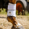 A close-up of a horse's lower leg and hoof wearing a white Woof Wear Club Brushing Boot outdoors on a gravel surface.