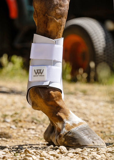 A close-up of a horse's lower leg and hoof wearing a white Woof Wear Club Brushing Boot outdoors on a gravel surface.