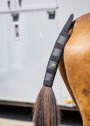 A close-up of a horse's tail wrapped in a Woof Wear Tail Guard with a small white logo patch, set against a blurred light gray trailer background.