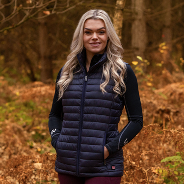 Woman wearing a navy heated puffer gilet standing in a forest with autumn foliage.